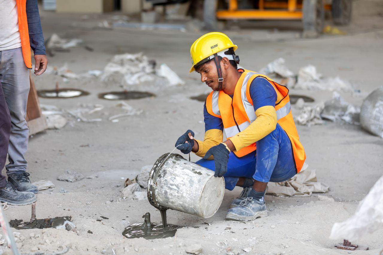 A construction worker kneels to pour concrete at a building site, wearing safety gear.